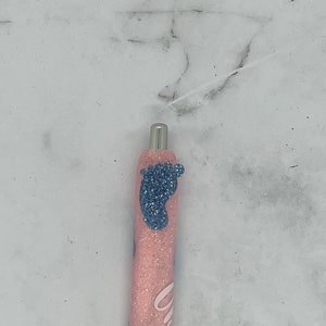 Op de afbeelding: Een blauwe en roze glitterpen met een baby-voetafdruk ontwerp. De pen heeft een zwarte inktpunt en een zilveren clip.