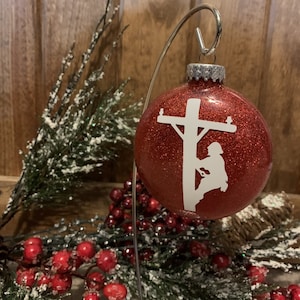 May include: Red glitter Christmas ornament with a white silhouette of a person working on a power line.