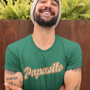 May include: A man wearing a green t-shirt with the text "Papasita" in a retro orange and yellow font. He is smiling and has a beard and tattoos.