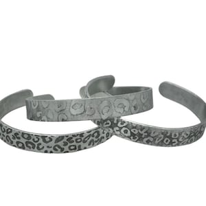 Può includere: Tre braccialetti a polsino argentati. Due hanno un motivo leopardato e uno ha un motivo floreale. I braccialetti sono aperti e sembrano essere in metallo. I braccialetti sono esposti su sfondo bianco.