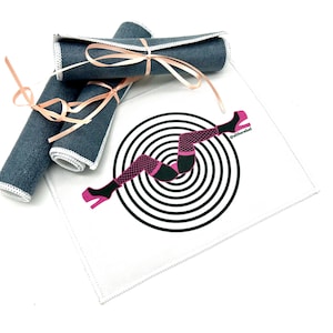 May include: A white fabric coaster with a black and white concentric circle design. A pink and black illustration of a woman's legs in high heels is on top of the circles. The text "@altherrebel" is printed on the coaster. Three rolled up blue fabric coasters are in the background.