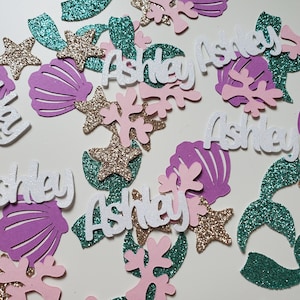 May include: A collection of mermaid-themed confetti featuring the name "Ashley" in white glitter letters, along with pink and purple seashells, green glitter mermaid tails, and gold glitter starfish.