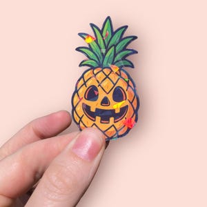 May include: A holographic sticker featuring a jack-o'-lantern pineapple design. The pineapple has an orange body with a carved face and green leafy top. The sticker has a glossy finish and is held against a light pink background.