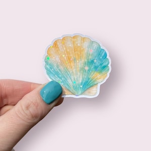 May include: A seashell sticker with a watercolor design. The sticker features a gradient of yellow, blue, and teal colors, with iridescent glitter accents. The sticker is held by a person with a teal fingernail.