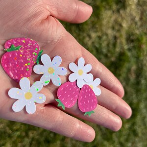 May include: A hand holding several paper cutouts of strawberries and daisies. The strawberries are pink with green stems and glitter accents. The daisies are white with yellow centers and glitter accents.