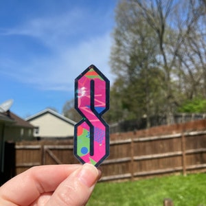 May include: A pink and black sticker with a stylized letter 'S' in a geometric pattern. The sticker has a glossy finish and is held in a hand.