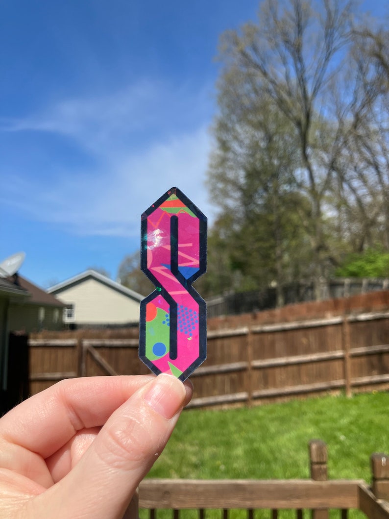 May include: A pink and black sticker with a stylized letter 'S' in a geometric pattern. The sticker has a glossy finish and is held in a hand.