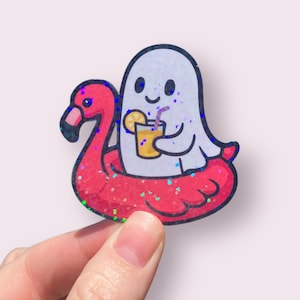May include: A sticker featuring a cartoon ghost floating on a red flamingo pool float. The ghost is holding a yellow drink with a straw and a lemon slice. The flamingo float has a glittery finish.