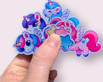 May include: A set of five iridescent holographic stickers featuring cartoon unicorns and stars. The stickers are blue, pink, and purple with glitter accents.