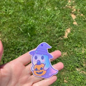 May include: A sticker featuring a cartoon ghost wearing a purple witch hat and holding an orange pumpkin. The ghost has a blue and purple color scheme with glitter accents. The sticker is held in a hand against a background of green grass.