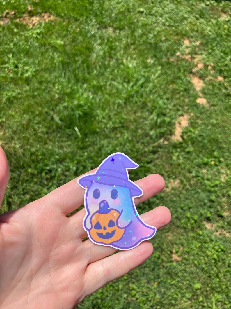 May include: A sticker featuring a cartoon ghost wearing a purple witch hat and holding an orange pumpkin. The ghost has a blue and purple color scheme with glitter accents. The sticker is held in a hand against a background of green grass.