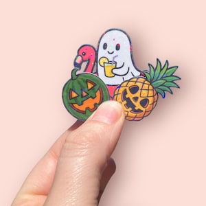 May include: A colorful sticker featuring a ghost holding a drink, a flamingo, a jack-o'-lantern, and a pineapple. The ghost is white with a smiling face. The flamingo is pink, and the jack-o'-lantern and pineapple have carved faces. The sticker is held by a hand.