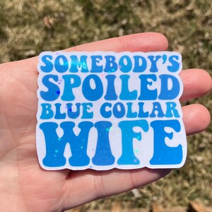 May include: A blue and white sticker with the text "Somebody's Spoiled Blue Collar Wife" in a retro font.
