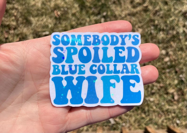May include: A blue and white sticker with the text "Somebody's Spoiled Blue Collar Wife" in a retro font.