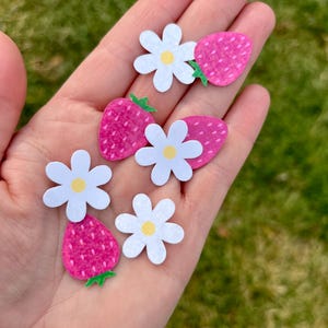 May include: A collection of handmade paper cutouts featuring pink strawberries and white daisies with yellow centers. The strawberries have green stems and textured surfaces. The cutouts are held in a hand against a blurred green background.