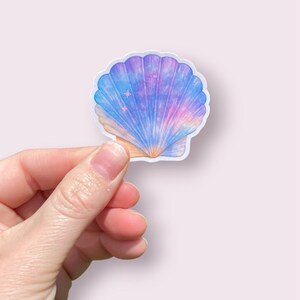 May include: A seashell sticker with a watercolor design. The shell is a light blue, purple, and pink color, with a few small star accents. The sticker is held by a hand against a light pink background.