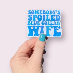 May include: A blue and white sticker with the text "Somebody's Spoiled Blue Collar Wife" in a retro font. The sticker has a glittery finish.