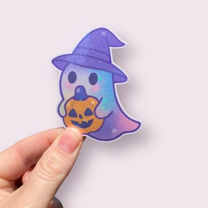 May include: A whimsical Halloween sticker featuring a cute ghost wearing a purple witch hat and holding an orange jack-o'-lantern. The ghost has a colorful, pastel body with glitter accents. A fun, festive design for Halloween.