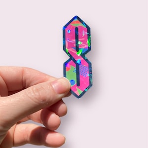 May include: A holographic sticker of the letter "S" in a stylized, old school font. The sticker is pink with a blue, green, and yellow geometric pattern.