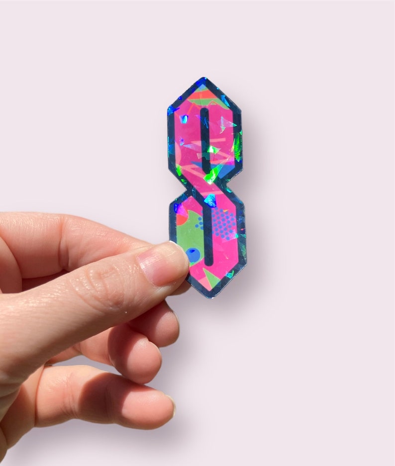 May include: A holographic sticker of the letter "S" in a stylized, old school font. The sticker is pink with a blue, green, and yellow geometric pattern.