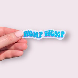 May include: A blue and white sticker with the text "WOMP WOMP" in a retro font. The sticker has a glittery finish.