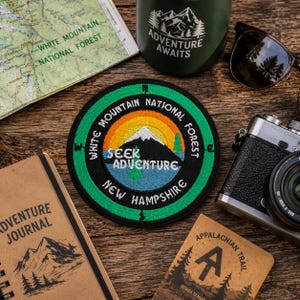 May include: A circular embroidered patch featuring a mountain landscape with a sun, trees, and the text "White Mountain National Forest New Hampshire" and "Seek Adventure" in white lettering, surrounded by a green border with compass directions.