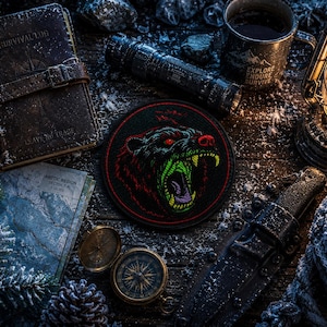 May include: A circular embroidered patch featuring a snarling bear head with red eyes and a green mouth, surrounded by a survival log, a tactical flashlight, a metal mug with text, a lantern, a map, a compass, a knife, and a rope on a dark surface.