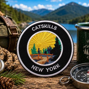 May include: A round black patch with the text "CATSKILLS NEW YORK" encircling a yellow sunburst design with trees and a bridge. A green enamel mug with "ADVENTURE AWAITS" is next to a compass and a backpack.