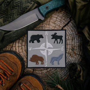 May include: A grey embroidered patch with a compass design and animal silhouettes: bear, moose, bison, and wolf. A large knife with a green handle is on a wooden surface, near brown hiking boots and a green scarf.