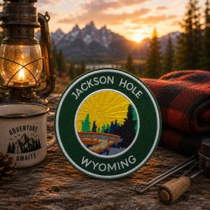 May include: A circular embroidered patch featuring a dark green border with the white text JACKSON HOLE WYOMING. The center depicts a yellow sun rising over a landscape with a wooden bridge, blue water, and dark green pine trees.