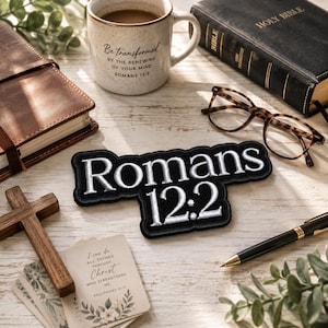 May include: A black embroidered patch with white text reading Romans 12:2, positioned among a ceramic mug with the same text, a black Holy Bible, brown-rimmed glasses, a wooden cross, a black pen, and a card with Philippians 4:13 text.