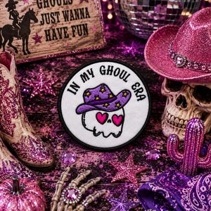 May include: A circular embroidered patch featuring a white skull with pink heart-shaped eyes wearing a purple cowboy hat with yellow stars. The text "IN MY GHOUL ERA" is stitched in black around the top edge of the white circular background.