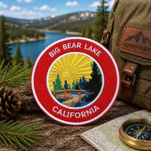 May include: A round, embroidered patch with a red border and the words "BIG BEAR LAKE CALIFORNIA" in white. The patch features a yellow sun, green trees, and a blue lake. A backpack, compass, and map are in the background.