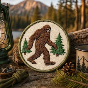 May include: Embroidered patch featuring a brown Bigfoot walking between two green pine trees. The patch has a cream-colored background with a green border. The patch is round and is set on a wooden surface with camping gear.