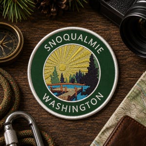 May include: Embroidered patch with a green border featuring the text "SNOQUALMIE WASHINGTON". The patch depicts a landscape with a sunburst, trees, and a lake. The patch is round and has a white border.