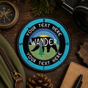 Op de afbeelding: Geborduurde patch met een berensilhouet en het woord "WANDER" in het wit. De patch heeft een blauwe en zwarte rand met de tekst "YOUR TEXT HERE". Een kompas, camera en andere outdoor items omringen de patch.