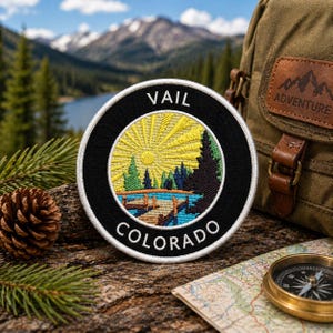 May include: Embroidered patch with a black border featuring the text "VAIL COLORADO." The patch depicts a yellow sun, green trees, and a blue lake. A brown backpack, a map, a compass, and a pinecone are also visible.