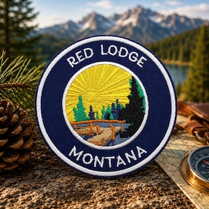 May include: Circular embroidered patch with the text "RED LODGE MONTANA". The patch features a landscape scene with a yellow sun, green trees, and a bridge, set against a blue background with a white border. The patch is on a textured surface.