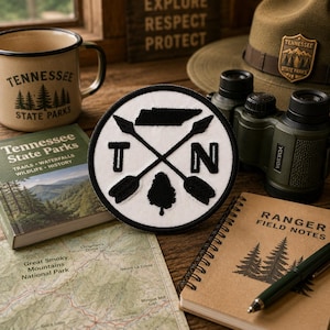 May include: A circular embroidered patch featuring a white background with a black border. The design includes a black silhouette of Tennessee, two crossed arrows, the letters T and N, and a black tree silhouette, all rendered in black thread.
