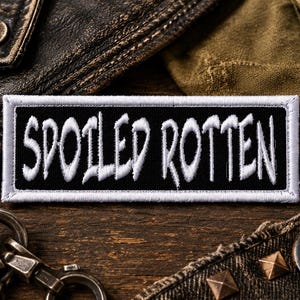 Könnte beinhalten: Ein rechteckiger schwarzer Aufnäher mit den Worten "SPOILED ROTTEN" in Weiß, umrandet von einem weißen Rand. Der Aufnäher ist vor einem Hintergrund aus Leder und Denim platziert, was auf eine Biker- oder Punk-Ästhetik hindeutet.