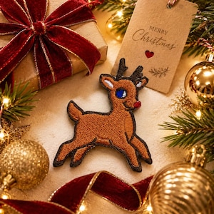May include: A brown embroidered reindeer patch featuring dark brown antlers, a bright red nose, and blue eyes. The patch is positioned centrally, surrounded by festive holiday decorations including a gift with a red velvet ribbon and a gift tag.