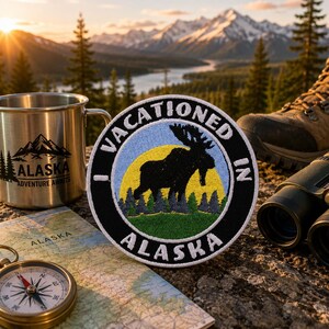 May include: A circular embroidered patch featuring a black silhouette of a moose against a yellow sun and blue sky, with green trees at the base. The black border contains the white text I VACATIONED IN ALASKA in a bold, sans-serif font.
