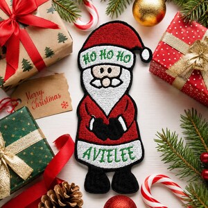 May include: An embroidered Santa Claus patch featuring a red hat with white trim, a white beard, a red coat with black gloves, and black boots. The text HO HO HO is stitched in green on the hat, and the name AVIELEE is stitched in green on the coat.