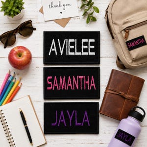 Peut inclure: Trois écussons rectangulaires noirs brodés avec les noms AVIELEE en blanc, SAMANTHA en rose et JAYLA en violet. Les écussons sont présentés avec un sac à dos beige, un carnet en cuir marron, une gourde violet clair et des articles de papeterie.