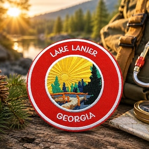 May include: A round, red Lake Lanier Georgia patch with white trim. The patch features a yellow sun, green and blue trees, and the text "Lake Lanier" and "Georgia". The patch is on a wooden surface with a backpack and compass in the background.