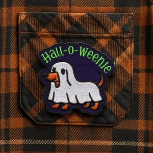 Peut inclure: Gros plan d'une poche de chemise à carreaux marron et noir avec un écusson sur le thème d'Halloween. L'écusson représente un chien fantôme blanc avec des détails orange et le texte "Hall-o-weenie" en vert.