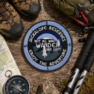 May include: A round, embroidered patch with the text "JACKALOPE RESERVES" and "NOT ALL WHO WANDER ARE LOST". The patch features a bear silhouette and a tree, with a light blue border. Hiking boots, a map, and a compass are also visible.