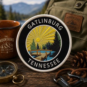 May include: Embroidered patch with a circular design featuring the text "GATLINBURG TENNESSEE". The patch depicts a sunburst over a lake and trees. The patch is black with white and yellow accents. Other items in the image include a compass, mug, and backpack.