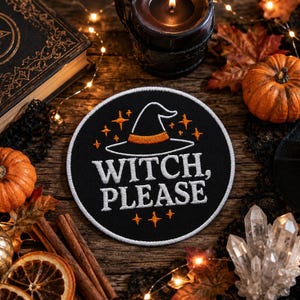 Pode incluir: Um emblema circular bordado em preto com um chapéu de bruxa branco com uma faixa laranja, o texto branco WITCH, PLEASE e pequenos detalhes de estrelas laranja. O emblema tem uma borda costurada branca sobre uma superfície de madeira escura.