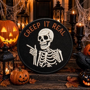 May include: A circular black embroidered patch featuring a white skeleton pointing with its right hand. The text "CREEP IT REAL" is stitched in orange thread in an arc above the skeleton's head. The patch has a black stitched border.